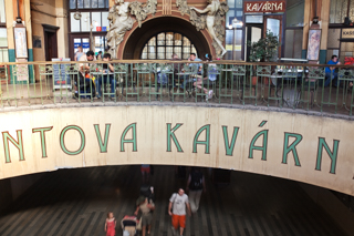 Bahnhof in Prag, zu sehen sind zwei Ebenen, unten laufen Menschen, oben sitzen Menschen in einem Cafe